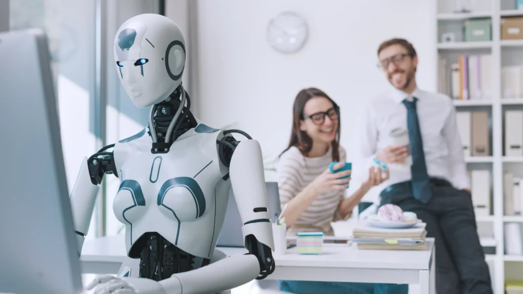 Humanoid robot working at a computer while two office colleagues talk and smile in the background
